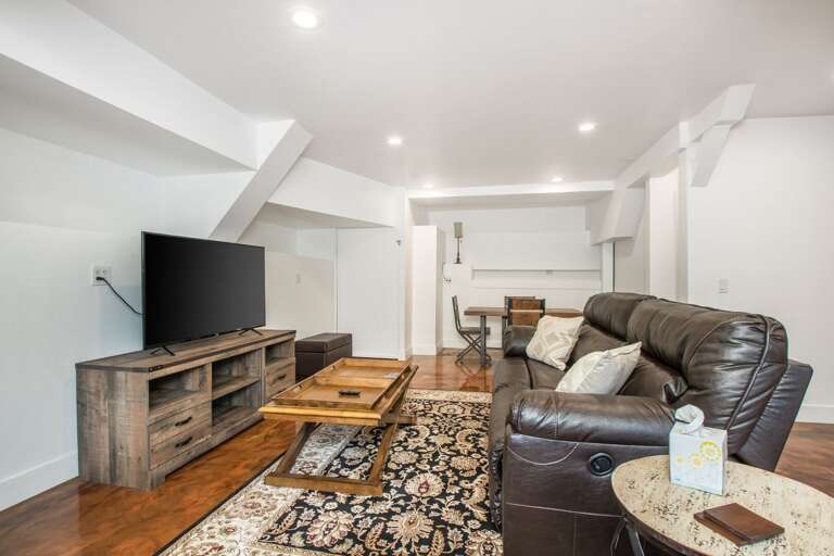 Located on the ground floor beneath the main house, this charming mother-in-law suite features polished epoxy floors, dark wood furnishings, a cozy leather sleeper-sofa, and a matching armchair.