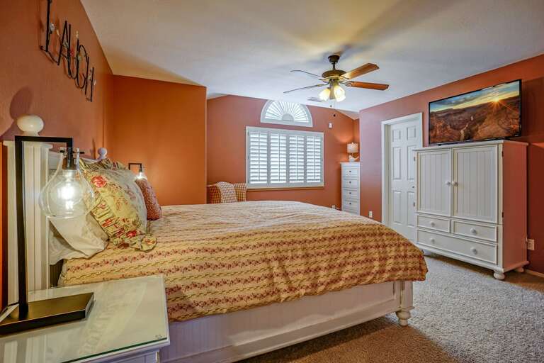 Upper Level: Bedroom 3 – Queen bed, ceiling fan, flat-screen TV atop a charming white armoire, and a cozy, inviting atmosphere. Upper Level: Bedroom 3 – Queen bed, ceiling fan, flat-screen TV atop a charming white armoire, and a cozy, inviting atmosphere.