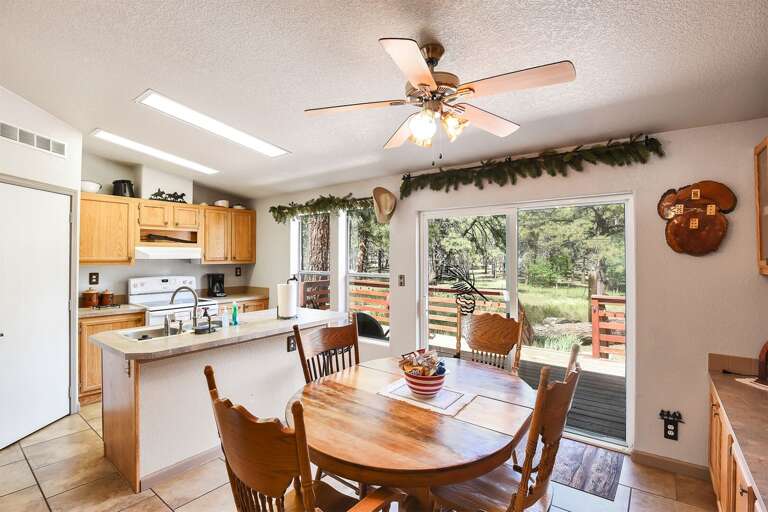 Bright, Breezy Dining Space With Wooden Chairs, Ceiling Fan, And Kitchen View, Leading To Leafy Outdoor Deck