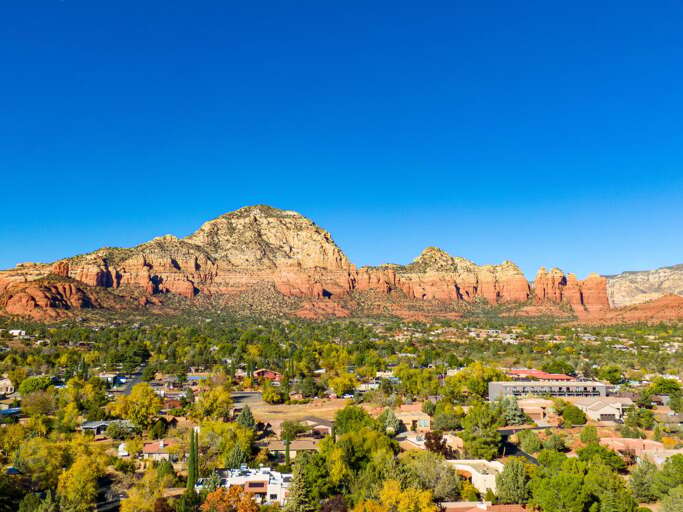 Aerial View Of A Vibrant Village Nestled Beneath Massive Red Rock Mountains