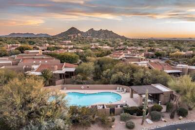 A serene desert community pool surrounded by lush greenery and adobe-style homes, with sweeping mountain views under a glowing sunset sky.