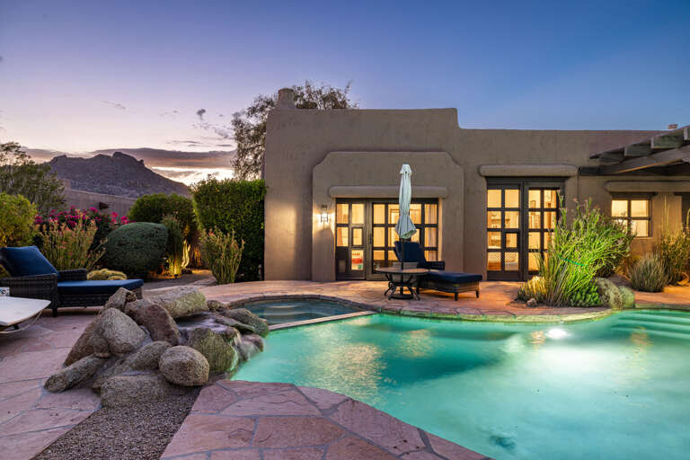 As the sun sets, the backyard transforms into a tranquil retreat with a glowing pool, ambient lighting, and beautiful desert surroundings.
