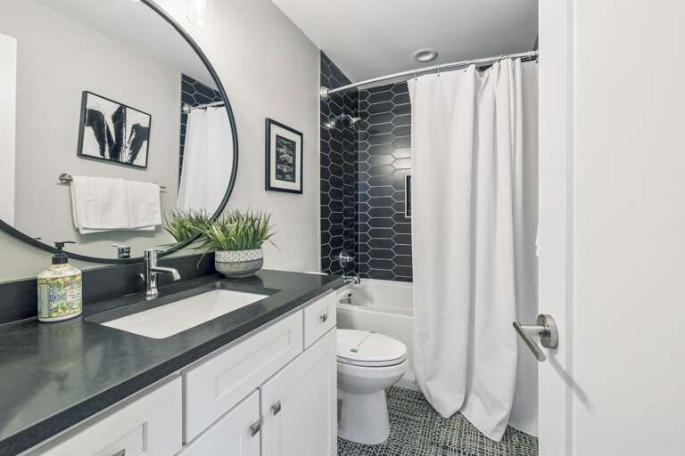 Striking black tile shower with geometric pattern and built in niche for essentials - Round mirror and dark vanity top add contrast and modern flair to the space - Bright lighting, framed art, and greenery finish the spa like look with style