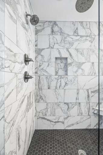 Frameless glass doors reveal stunning marble tile with soft gray veining throughout - Built in niche for convenience and multiple spray heads for a refreshing experience - Soft lighting and stone style flooring create a calming, upscale shower retreat