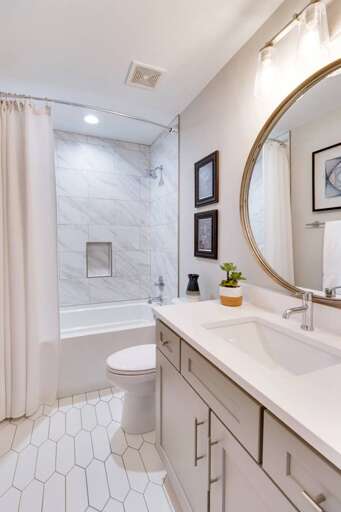 - White vanity with quartz top and brushed nickel fixtures offers a spa like feel - Full tub with marble style tile and inset shelf perfect for a relaxing soak - Round mirror, framed art, and fresh greenery complete the calming design  