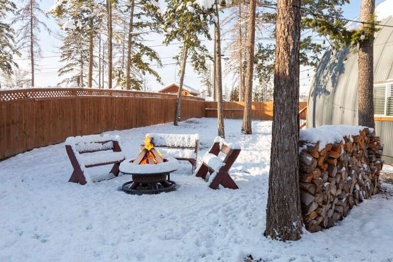 Snowy Scene With Seating Around Small Fire, Fence And Trees, Firewood Stacked