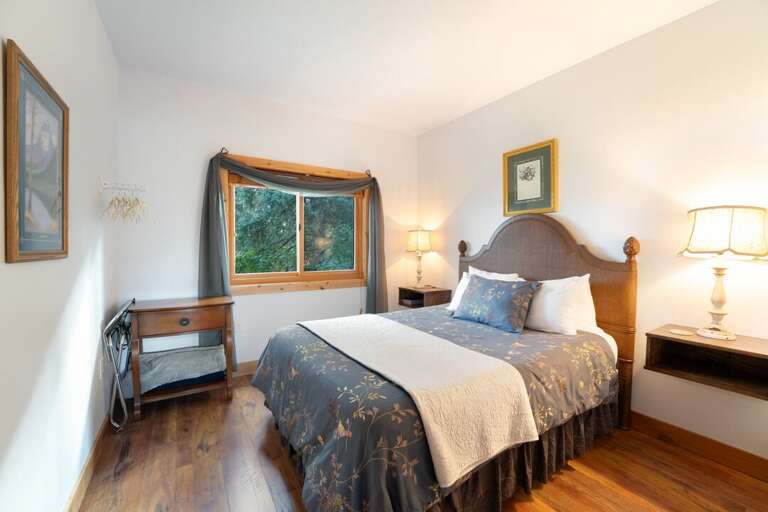 A cozy second bedroom featuring warm lighting with a window overlooking the surrounding trees. | Main Level