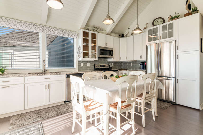 White Kitchen With Wooden Chairs Around A Dining Table, Black Tiled Backsplash Behind Stove, Bright Light Filtering Through Window Blinds