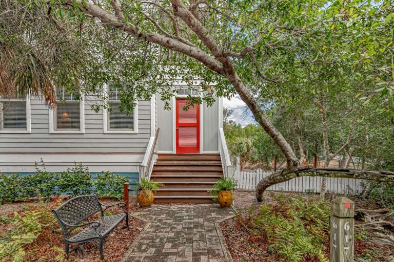 Entry Path Leading To A Red Door Flanked By Lush Foliage And A Sitting Bench Under An Arching Tree