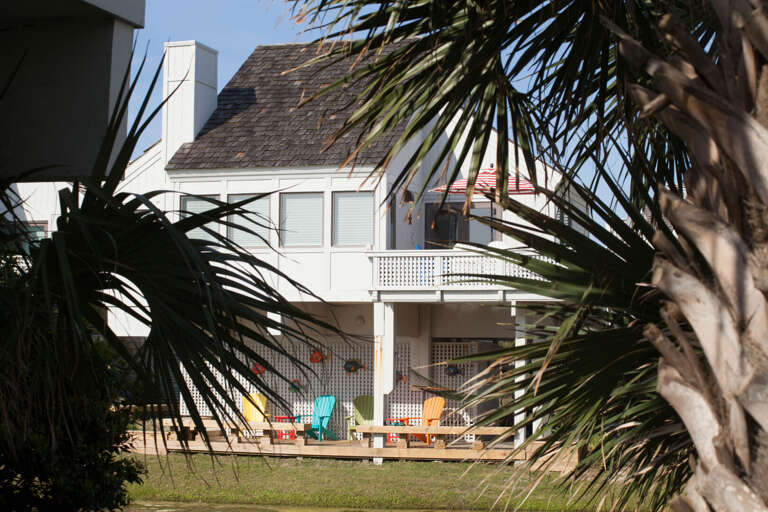 White Building Between Palm Branches With Colorful Chairs On Grass