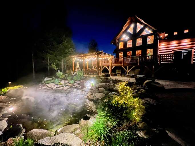 Illuminated Cabin At Night With Pond And Landscaping