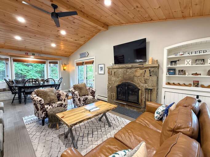 Gather in the main-level living room, featuring a stone fireplace, TV, leather sofa, and warm wood ceiling.