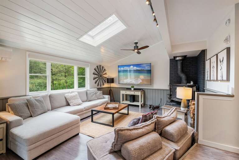 The TV and woodstove face generous seating beneath skylights and wide woodland windows.