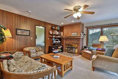 Living area in warm wood and beige tones, with seating, a Smart TV, fireplace, coffee table, ceiling fan, and a large window framing mountain views.