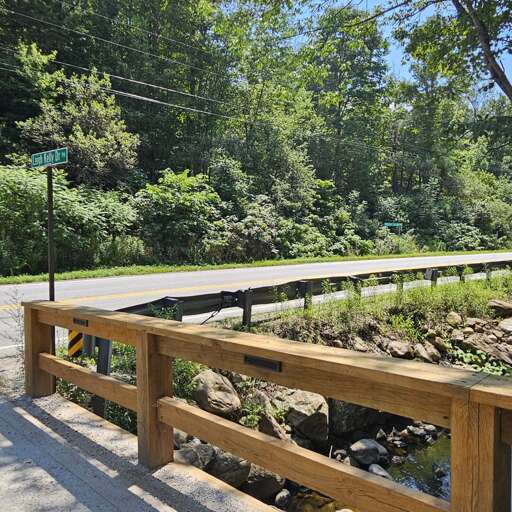 The wooden bridge crosses a rocky creek beside the tree-lined road.