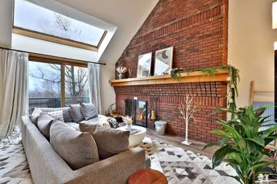 Cozy living room with a large L-shaped sofa, a balcony and a high window showing mountain views, and an exposed-brick wall with a fireplace.