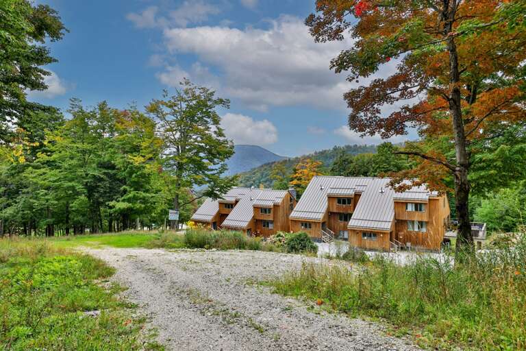 A wooded ridge rises beyond the condo buildings, with the gravel approach winding through the trees.