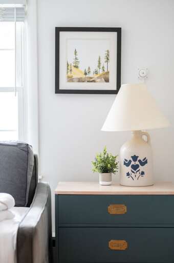 Bedroom nightstand with a lamp, framed artwork, and small plant beside the bed.