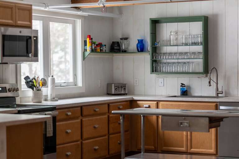 Kitchen sink area with wood cabinetry, dishwasher, window, and wall-mounted glass storage.