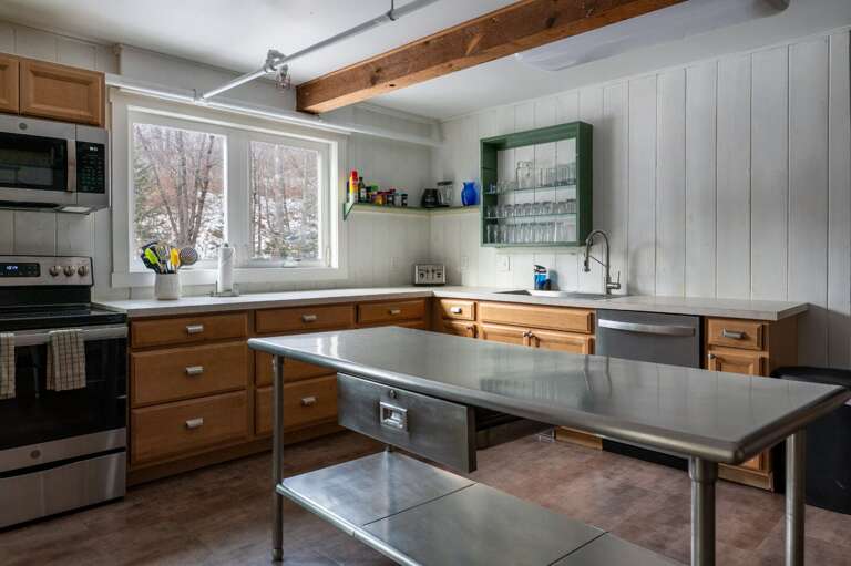 Kitchen with a stainless steel prep table, wood cabinetry, dishwasher, range, and window over the sink.