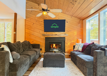 Two sofas face the fireplace beneath the tall wood ceiling, with broad windows looking out toward the trees.