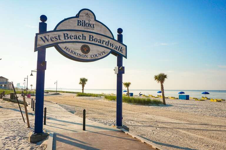 Biloxi West Beach Boardwalk Sign With Sandy Beach And Clear Sky In Background Biloxi West Beach Boardwalk Sign With Sandy Beach And Clear Sky In Background