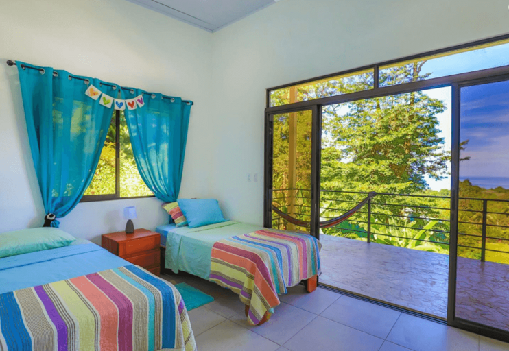 Bright Room With Two Beds, Blue Curtains, And Sliding Door Leading To Balcony With Greenery View