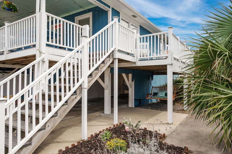 Blue Beachside Building With White Wooden Stairs And Balcony, Surrounded By Sand And Shrubs