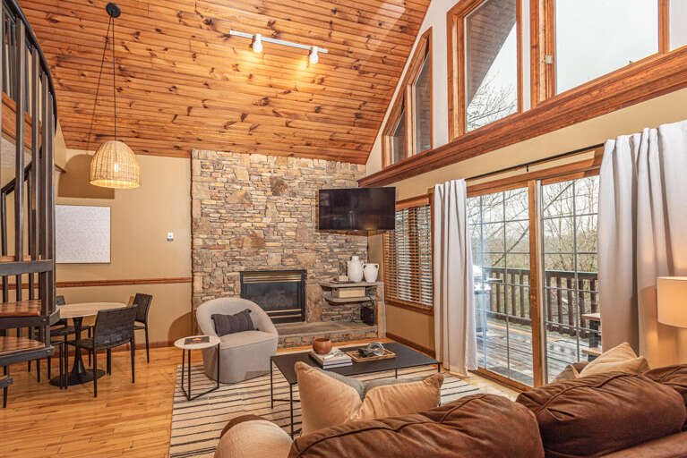 Spacious Living Space With Stone Fireplace, High Wooden Ceilings, And Large Windows Overlooking Leafy Locale