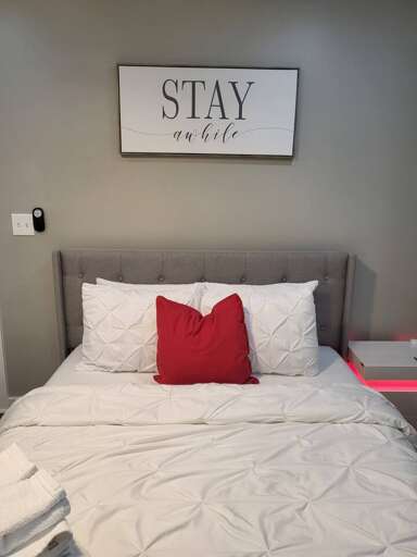 Bed With White Bedding, Red Pillow, And STAY Awhile Sign Above