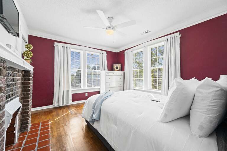 The bedroom boasts rich red walls and a cozy white bed adorned with plush pillows and a light blue throw, adding a touch of elegance. A striking brick fireplace serves as the focal point, complemented by white linens The bedroom boasts rich red walls and a cozy white bed adorned with plush pillows and a light blue throw, adding a touch of elegance. A striking brick fireplace serves as the focal point, complemented by white linens