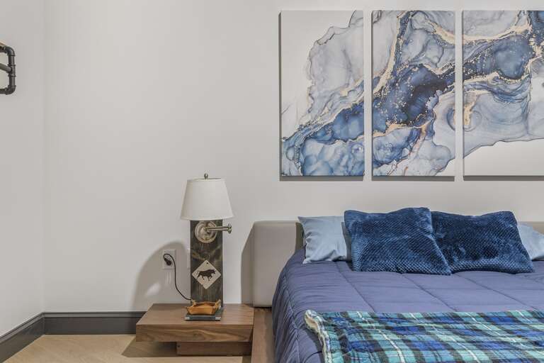 Close-up of the bedroom showcasing a detailed art piece above the bed, adding a splash of color and elegance to the room, accompanied by a rustic bedside lamp. Close-up of the bedroom showcasing a detailed art piece above the bed, adding a splash of color and elegance to the room, accompanied by a rustic bedside lamp.