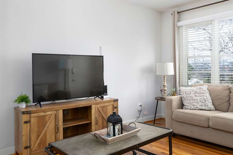 A bright and inviting living room with a large smart TV, cozy beige couches, and plenty of natural light streaming through the windows. Perfect for relaxation or movie nights. A bright and inviting living room with a large smart TV, cozy beige couches, and plenty of natural light streaming through the windows. Perfect for relaxation or movie nights.