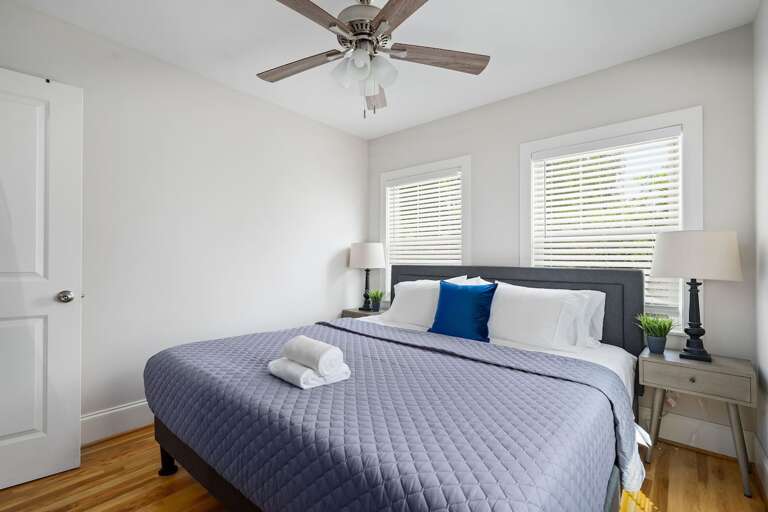 This master bedroom offers a king-sized bed set in a calming color scheme, complete with ceiling fan and ample natural light for a peaceful retreat.