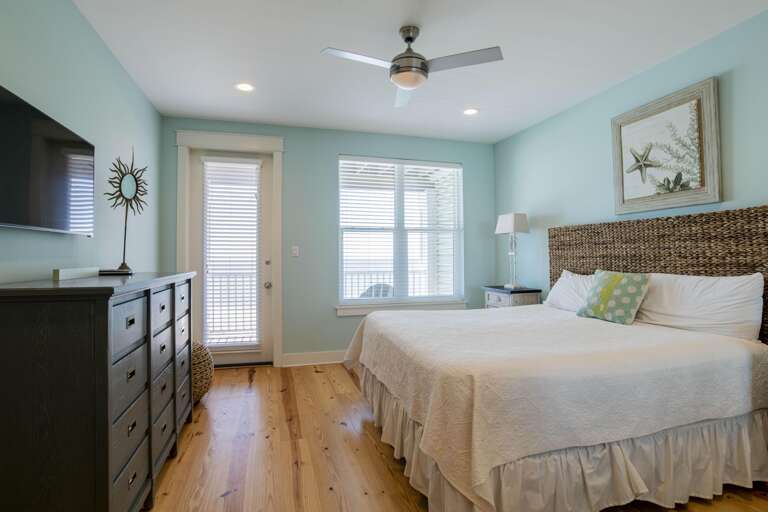 Light pine floors stretch beneath a white quilted bed and lead to a glass door and window with white blinds.