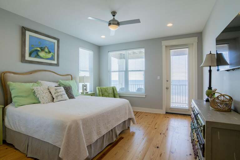 Light wood floors extend beneath a beige upholstered headboard, green accent chair, and a colorful multi-drawer dresser.