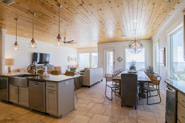 Open-plan living area with limestone tile flooring, wood-paneled ceiling, and glass pendant lights over the kitchen island.
