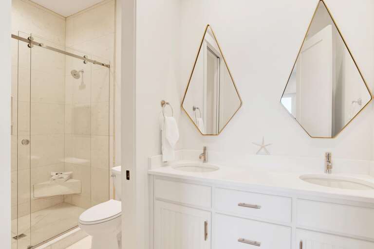 Dual sinks set in white quartz countertop with diamond-shaped mirrors above and glass-enclosed tiled shower with built-in bench.