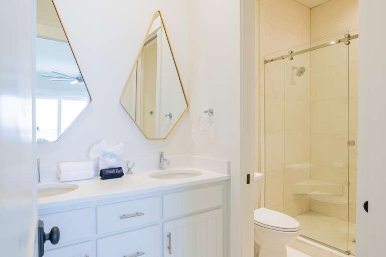 Bathroom with dual white vanities, diamond-shaped mirrors, and a glass-enclosed beige tiled shower with corner bench.