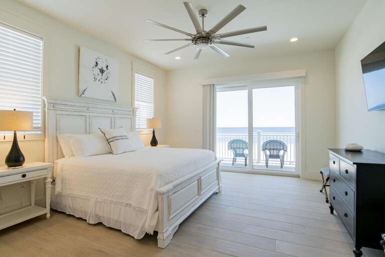 King bed with white distressed wood frame, matching nightstands, and sliding glass doors opening to balcony with ocean view.