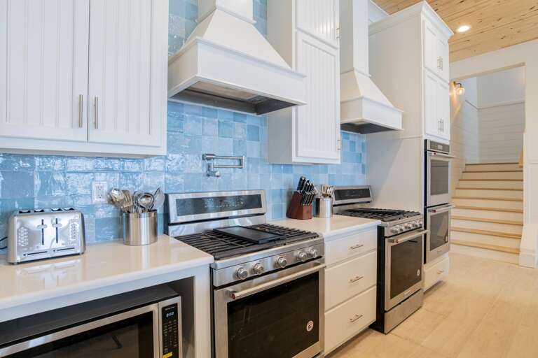 Dual stainless steel gas ranges sit beneath white vent hoods and glossy blue tile backsplash.