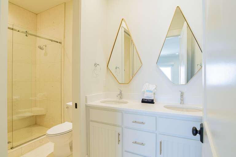 Bathroom with dual white vanities, diamond-shaped mirrors, and a glass-enclosed beige tiled shower.