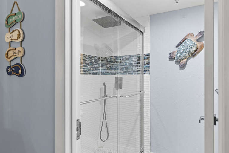 A bright walk-in shower featuring a sleek glass door and blue mosaic tile accents. Beach-themed decor enhances the serene coastal feel of this refreshing space.
