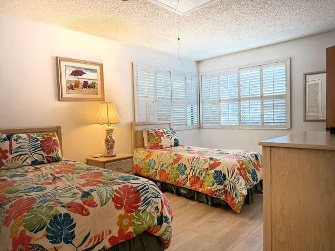Perfect for a beach stay, this cozy bedroom features two twin beds with tropical bedding, natural light through plantation shutters, and cheerful coastal décor for a relaxing retreat.