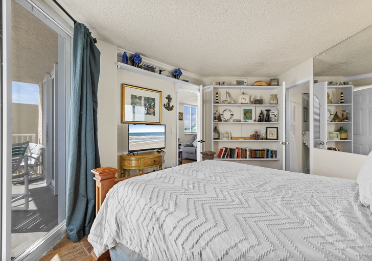 Wake up to ocean views in the master bedroom featuring balcony access, a flat-screen TV, and built-in shelving with coastal décor—perfect for a relaxing beach stay retreat.