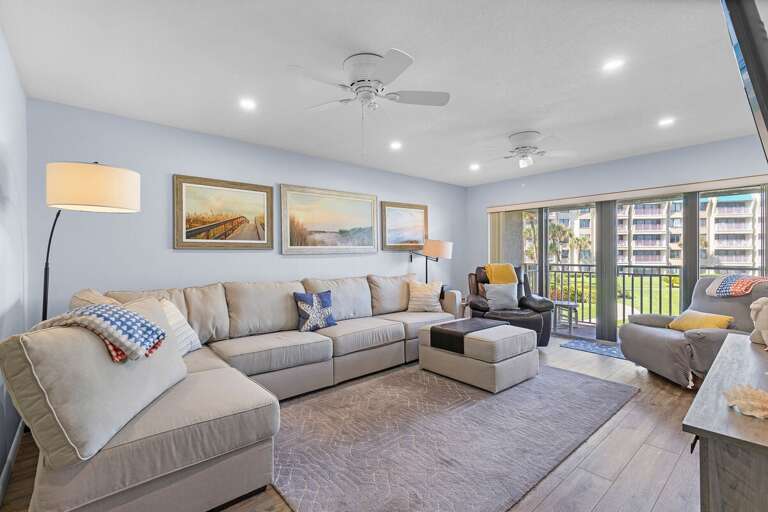 Enjoy ocean breezes in this bright, beach-inspired living room featuring a plush sectional, coastal décor, and large windows opening to serene balcony and resort views.