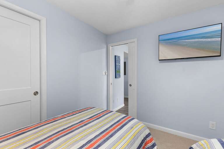 This inviting beach-themed guest room features twin beds with colorful linens and a wall-mounted TV—perfect for relaxing after a day of seaside fun or catching a sunset movie night.
