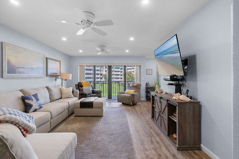 Relax in this spacious beach-inspired living room featuring plush seating, a large TV, and balcony access with views of the ocean breeze and tropical landscape. Perfect for your coastal getaway.