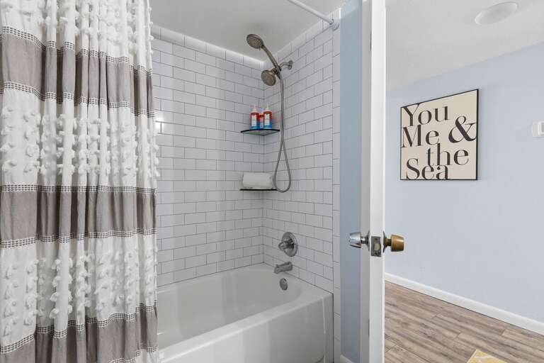 Unwind after your beach day in this inviting bath featuring a sleek tiled shower-tub combo, coastal décor, and plush towels—perfect for a refreshing rinse or a soothing soak by the sea.