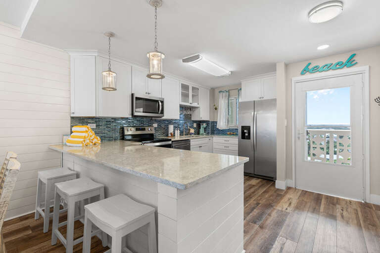 Bright beachside kitchen featuring stainless appliances, ocean-inspired backsplash, and bar seating. Enjoy modern coastal style and easy access to stunning seaside views.